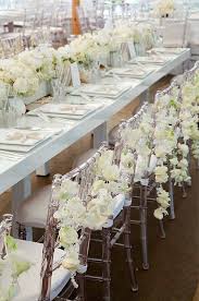 Love The Flowers On The Back Of The Chairs Clear Chiavari Chairs Are Decorated With Garlands Of Reception Table Linens All White Wedding Wedding Chairs