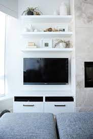 Gray Stools Face Three Styled White Floating Shelves Fixed Above A Wall Mounted Flat Panel Television Posi Wall Mounted Tv Shelves Above Tv Wall Mount Tv Ideas