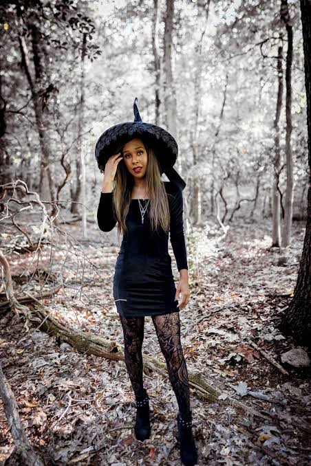 Concise (Best for Accessibility)

Woman posing in the woods wearing a classic black witch costume with a wide-brim pointed hat, long-sleeved mini dress, patterned lace tights, and black boots.