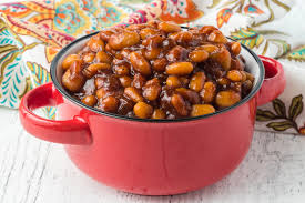 A Red Pot Filled With Baked Beans On A White Wooden Surface, With A Colorful Patterned Cloth In The Background.