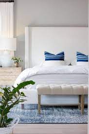 White Wingback Bed Displaying Blue Striped Pillows White Bedding And An Ivory Tufted Bench At The Foot Of White Upholstered Bed Upholstered Beds Wingback Bed