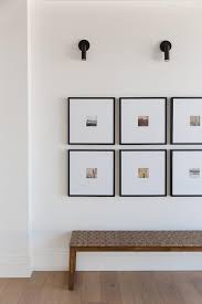 Stacked Picture Gallery Frames Are Lit By Matte Black Picture Lights And Mounted Over A Wood And Fabric Gallery Wall Picture Frame Gallery Entryway Decor Small
