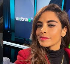 Apresentadora de TV sorri para a câmera em frente a um logo "extras" em um estúdio.