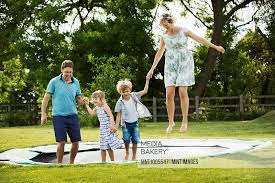 Mediabakery - Photo by Mint Images - Man, woman, boy and girl holding  hands, jumping on a trampoline set in the lawn in a garden.