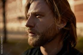 Close up image of fashionable young Caucasian man with long hair and ginger  beard posing outdoors wearing earring and facial piercing, having serious  look, squinting eyes because of sunshine Stock Photo |
