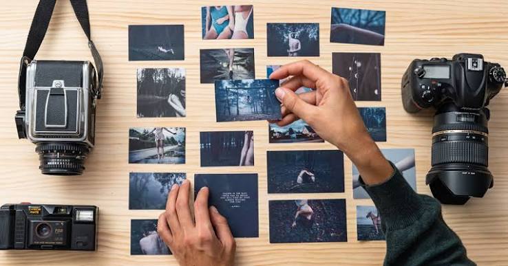 "Fotografia no estilo flat lay (vista de cima) sobre uma superfície de madeira clara. No centro, duas mãos organizam cuidadosamente várias fotografias impressas de pequeno formato, com tom azulado e atmosfera melancólica, retratando paisagens e figuras humanas. À esquerda, há duas câmeras fotográficas vintage (uma de médio formato e uma compacta). À direita, encontra-se uma câmera DSLR moderna preta com uma lente grande."