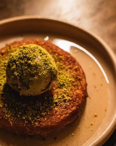 Pouring cooled sugar syrup over freshly baked hot kunefe in a pan, creating steam and crispness