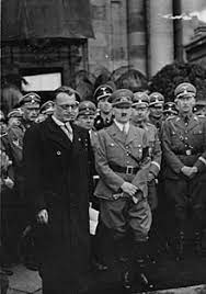 Black and white photograph of a man in glasses addressing a crowd of men in suits, with guards in uniform behind them. Arthur Seyss Inquart Wikipedia