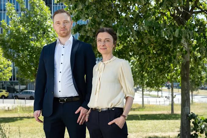 A man and a woman in business casual attire stand outdoors near trees and a modern office building.
