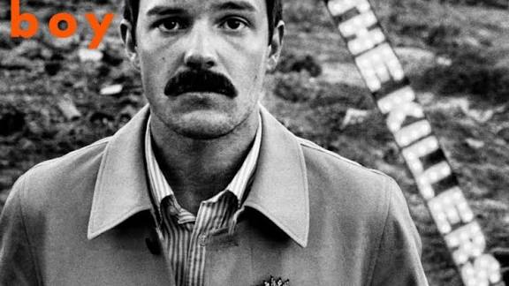 Black and white film photograph of a man with a distinctive mustache wearing a collared shirt and light jacket, shot against a blurred outdoor background with a ladder visible behind him.