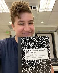 ID: The first image is a selfie of Ace in a Caltech classroom. They're  wearing a dark blue t-shirt and holding a black-and-white composition  notebook. They wrote “NITARP Notebook, Ace Schwarz (they/them)