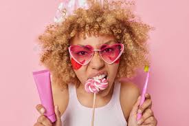 Dissatisfied young woman with curly hair keeps caramel candy in mouth holds  toothpaste and toothbrus Stock Photo by wayhomestudioo