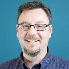A smiling man with glasses and a goatee wearing a checkered shirt against a  blue background.