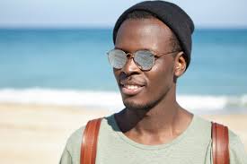 Retrato de un alegre viajero de hombre negro que disfruta de las vacaciones  de verano junto al mar, con aspecto despreocupado y relajado, con sombrero  de moda y gafas de sol con