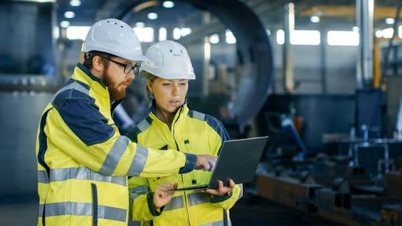 Dois profissionais com capacete e colete de alta visibilidade a consultar um computador em ambiente industrial