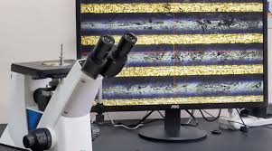 도립형 금속현미경의 스테이지 위에 연마된 시편이 놓여 있고, 모니터 화면에 복잡한 금속 결정립 구조가 확대되어 나타난 모습