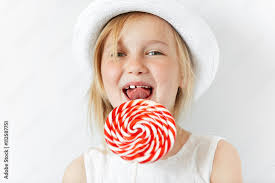 Nice little girl smiling at camera in white morning room, ready to lick red  round lollipop. Joyful atmosphere and happy child with yummy candy is a way  to a good mood. Stock