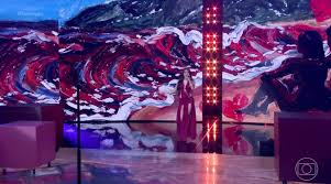 Cantora em palco com painel de LED exibindo arte abstrata de ondas vermelhas e azuis. Ao fundo, silhuetas de pessoas sentada…