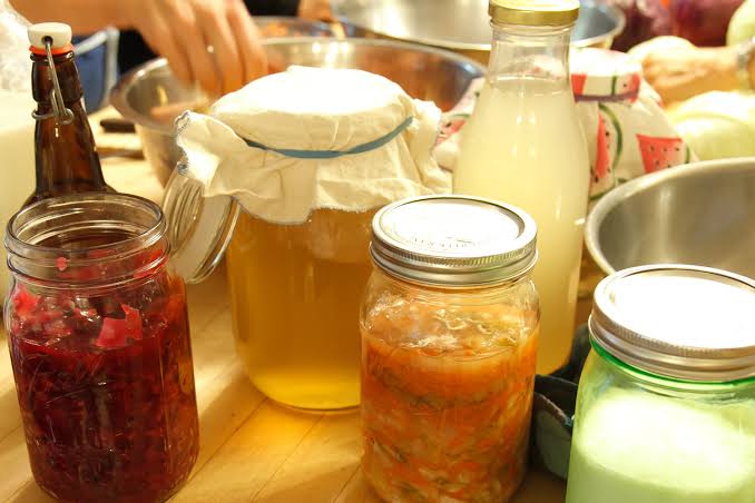 A person stirring a glass jar of homemade fermented vegetables with a wooden spoon in a bright kitchen