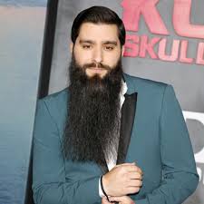 Handsome bearded man with long beard and mustache has stylish hair on surprised face holding glass of alcoholic beverage in vintage suede leather waistcoat o white brick wall studio background. How To Grow A Beard How To Trim A Beard For Your Face Shape