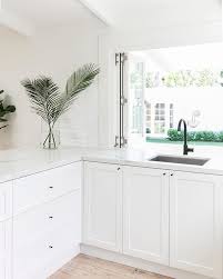 Country style kitchen with shaker cabinets accented with latch and cup pull hardware below a white marble counter with matching marble backsplash and glass front upper cabinets with blue interiors to the left of the farmhouse sink with hook spout faucet next to a stainless steel dishwasher. 12 Best White Kitchen Cabinets Black Hinges And Hardware Ideas White Kitchen Cabinets New Kitchen Kitchen Design