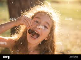 A cheerful sweet girl with cherry berries in her mouth. Funny summer  portrait of a child with a cherry, gifts of summer, summer time Stock Photo 