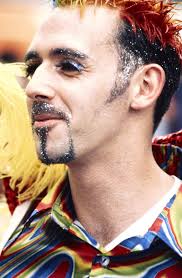 Context: Close up of Eddie McGuinness in colourful shirt, glitter make-up,  and rainbow coloured hair.