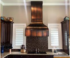Brass rivet straps accent a gray wooden range hood above an integrated gas cooktop in a gray and gold transitional kitchen. Custom Copper Stainless Range Hoods Havens Luxury Metals
