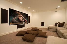 Chic basement movie room features a gray ceiling over walls clad in gold grasscloth paneled walls framing a long light taupe sofa under a ceiling mounted movie projector placed in raised platform facing additional seating in the form of a light taupe slipcovered sectional flanked by wood and iron end tables alongside three moroccan leather poufs atop a light taupe geometric rug facing a large. Basement Home Theater Design Ideas For Your Modern Home