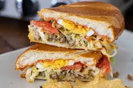 A closeup of a burger patty on a bun with lettuce, tomato, melted cheddar, and caramelized onions Dominican Riki Taki Belqui S Twist