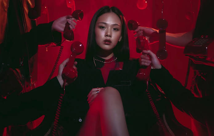 A dramatic cinematic close-up of South Korean singer BIBI in a scene from "The Weekend" music video. The shot is heavily saturated in intense crimson and deep red ambient lighting, casting dark shadows that enhance the eerie atmosphere. Her long dark hair is pulled back, framing her face as she stares directly into the camera with a piercing, supernatural expression. Dark, veiny black lines crawl outwards from her nose and cheeks across her pale skin, and her irises glow faintly with an unsettling, otherworldly light, capturing her visual transformation into a vampire-like creature.
