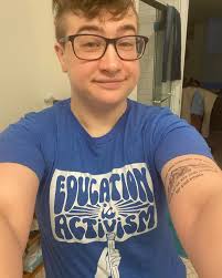 ID: A selfie of Ace wearing a @kindcotton t-shirt. The shirt is a darkish  blue with a white image and text in the center. A hand is holding a pencil  like a