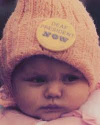 One of the youngest marchers wears a Deaf President Now button. Color  closeup photo of a baby looking at the distance wearing a pink beanie with  the button