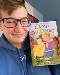 ID: A selfie of Ace grinning broadly and holding up the book Camp QUILTBAG  by Nicole Melleby and A.J. Sass. Two middle-school aged students are  standing together in the center. One with