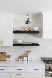 Beside White Kitchen Cabinets Adorning Brass Hardware Two Stacked Dark Stained Floating Shelves Are Lit By A Clear G Home Kitchens Kitchen Design Chic Kitchen