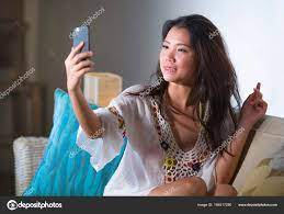 年轻美丽和快乐的亚洲华人20s 或30s 女人拿着手机自拍照片坐在家里客厅沙发沙发上微笑着—  图库照片©TheVisualsYouNeed＃194017290