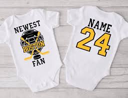 two white Bruins onesies; one says "newest Boston fan" in yellow in front of a black hockey helmet and crossed sticks"; the second is the back of the jersey with a large yellow and black number 24