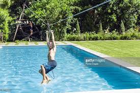 Full Length Of Boy Hanging From Zip Line Over Swimming Pool Pool Swimming Pools Zip Line Backyard