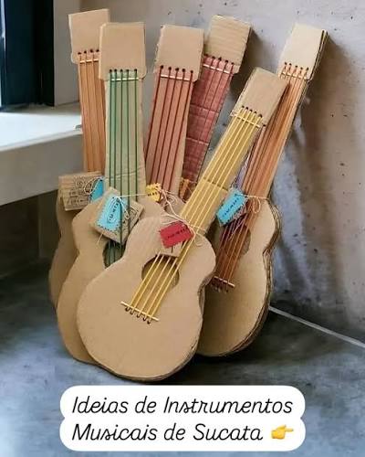 na imagem tem cinco violões feitos de papelão sobre o chão de cimento, encostados numa parede lisa com bege.
