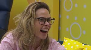 A mulher de cabelos loiros e óculos, sorrindo com a boca aberta, em um ambiente com fundo amarelo e detalhes circulares.