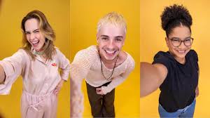 Três pessoas sorrindo para a câmera em um fundo amarelo. Da esquerda para a direita: uma mulher loira, um homem com cabelo l…