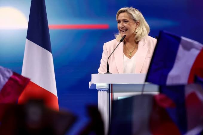 Marine Le Pen speaking at a podium with the French flag in the background.