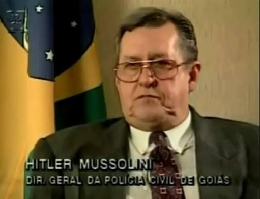 A man in his fourties, wearing a suit and glasses stands before a white background and a Brazilian flag. The image is subtitled with his mispelt name and title: Hitler Mussolini, Goiás's Municipal Police Director.