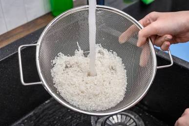 Uncooked white rice grains being rinsed in a fine mesh strainer under a kitchen tap running water