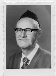 A portrait of Rabbi Yizhak Zvi Ettinger, with a smile and a tall black  kippah, and wearing a suit and tie