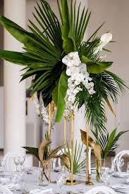 A Glam Tropical Centerpiece Of Large Tropical Leaves And White Orchids On Tall Tropical Wedding Centerpieces Flower Centerpieces Wedding Tropical Centerpieces