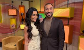 Apresentadores Patrícia Poeta e Manoel Soares sorriem em estúdio de TV com fundo colorido e cadeiras.