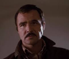 Which Burt Reynolds movie features a scene where he hides in a small  building and punches a guy through a window?