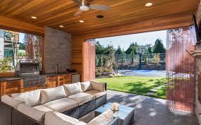 A gas grill with granite countertops and custom cabinetry is framed by large stone pillars that also frame the view to the garden and support a large wood roof covering. How To Build An Outdoor Kitchen