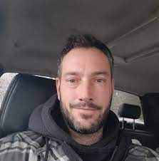 A person with a beard is sitting in a car, smiling slightly.